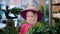 Young cute girl in hat and pink jacket looking green plants in orangery. Florist woman considering plats in indoor