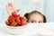 Young cute smiling european little girl is trying to steal ripe jucy strawberry from plate of many berries while she