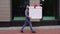 Young delivery man walking along city street, carrying in hands surprising festive cardboard box.