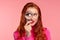 Young detective. Close-up portrait of surprised redhead girl looking at camera through magnifying glass, over pink