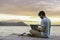 Young digital nomad man sitting on wooden pier at sea working on internet remotely at sunset - Traveling with a computer - Online