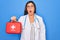 Young doctor woman holding medical first aid kit red box for emergency over blue background scared in shock with a surprise face,