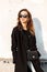 Young elegant hipster woman in trendy sunglasses in a long black coat in jeans with a handbag near a white building enjoys