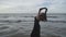 Young elegant woman in black long dress dancing with hands up holding black scarf on the pier