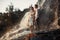 Young enamored couple stands on rock under spray of waterfall.