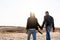 Young European couple dressed leather jacket and blue jeans walking desert holding hands view from the back