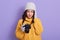 Young European female against lilac wall, photographer woman holding camera and keeping palm on her head, looks nervous and very