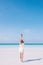 A young European long hair woman stands on a white sandy beach by the ocean. A girl in a white dress is holding a pineapple.