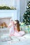 Young european mother holding daughter in arms near decorated fireplace and Christmas tree, wearing pink dress.