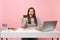 Young excited woman holding credit card while sit work at white desk with contemporary pc laptop isolated on pastel pink