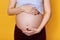 Young faceless pregnant woman holds her big belly with hand isolated over yellow background. Pregnant model being photographed in