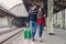 Young family couple embrace and look at each other with smile, wear jeans and warm sweaters, carry suitcase, pose on railways