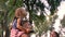 Young family is walking in park in summer, girl sits on father`s shoulders and they watch up at trees