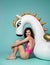 Young fashion woman relaxing with big inflatable unicorn pegasus float mattress in pink bikini for swimming pool