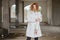 Young fashionably dressed red-haired girl with curly hair in a white coat posing, looking at the camera in an abandoned room