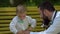 A young father with a beard sits on a yellow bench in the park and takes pictures of his young son.