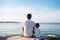 Young father with his son wearing white t-shirt sitting on shore of sea or ocean. Rear view. Mock up template for t