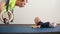 Young father pressed on the gymnastic rings. Next to him lie a little baby plays with a toy. Man kisses the child`s