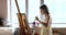 Young female artist drawing standing near easel in creative workshop