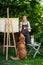 Young female artist working on her art canvas painting outdoors in her garden with golden retriever keeping her company.