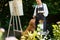 Young female artist working on her art canvas painting outdoors in her garden with golden retriever keeping her company.