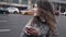 Young female city dweller is using smartphone standing near road in town, viewing social nets and apps