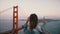 Young female creative worker with backpack walks watching epic scenery, famous sunset Golden Gate Bridge in strong wind.