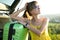 Young female driver having rest leaning on a green suitcase near her car in summer nature. Travel and vacations concept