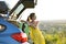 Young female driver having rest leaning on a green suitcase near her car in summer nature. Travel and vacations concept