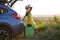 Young female driver having rest sitting on a suitcase near her car in summer field. Travel and vacations concept