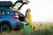 Young female driver having rest sitting on a suitcase near her car in summer field. Travel and vacations concept