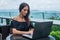 Young female freelancer dressed in black working on a project laptop computer while sitting at rooftop cafe terrace