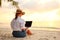 Young female freelancer wearing straw hat working on laptop while sitting on tropical beach at sunset