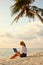 Young female freelancer wearing straw hat working on laptop while sitting on tropical beach at sunset