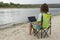 Young female with laptop in her knees is sitting camping chair on the bank of picturesque river.