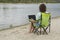 Young female with laptop in her knees is sitting camping chair on the bank of picturesque river.