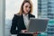 Young female lawyer working in her luxurious office holding a laptop standing against panoramic window with a view on