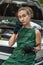 Young female mechanic in green uniform standing near a broken-down car and waiting help