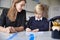 Young female primary school teacher and schoolboy sitting at a table working one on one, looking down, front view, close up