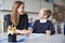 Young female primary school teacher and schoolboy sitting at a table working one on one, looking at each other smiling, front view