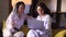 Young female professional working on computer device. Sitting on sofa with headphones, her friend - stylish woman with