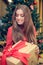 Young female is sitting in front of the christmas tree and have gift box with red ribbon in her hands