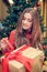 Young female is sitting in front of the christmas tree and have gift box with red ribbon in her hands