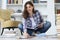 Young female student studying at home, sitting on floor against cozy domestic interior, surrounded with pile of books