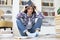 Young female student studying at home, sitting on floor against cozy domestic interior, surrounded with pile of books