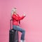 Young female tourist in a jacket and with Luggage on a pink background.