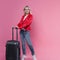 Young female tourist in a jacket and with Luggage stands on a pink background.