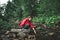 Young female tourist in a red raincoat touches the mountain stream with her hand, copy space. Hiker girl in a red jacket crouches