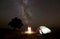 Young female tourist resting near illuminated tent, camping in mountains at night under starry sky