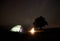 Young female tourist resting near illuminated tent, camping in mountains at night under starry sky